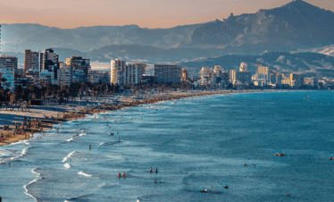 Panoramic view of Benidorm coastline, showing a crowded sandy beach, turquoise Mediterranean waters, high-rise buildings along the shore, and mountains in the background in Spain.