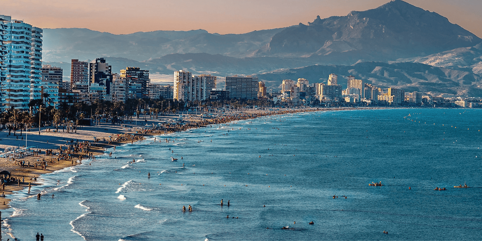Panoramic view of Benidorm coastline, showing a crowded sandy beach, turquoise Mediterranean waters, high-rise buildings along the shore, and mountains in the background in Spain.