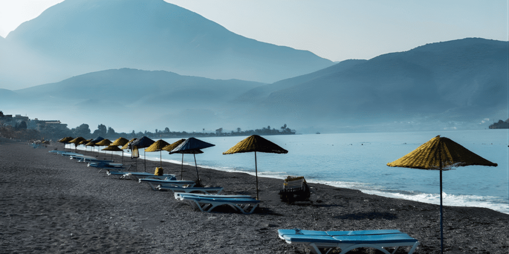 Pebble beach with sun loungers and colorful umbrellas along a calm sea with mountains in the background