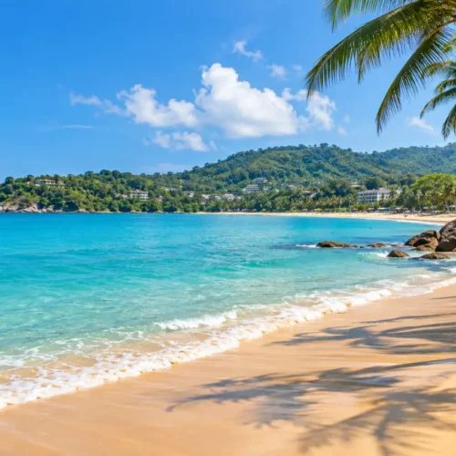 Scenic Phuket beach with turquoise water, golden sand, palm trees and lush green hills under a clear blue sky