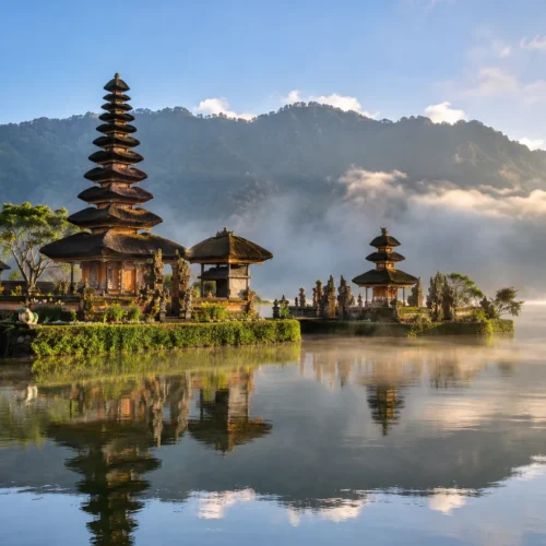 Traditional Balinese lake temple with multi-tiered pagoda roofs reflected in calm water, surrounded by misty mountains and tropical greenery.