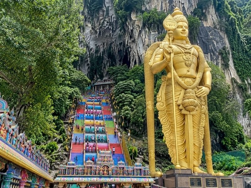 Golden Lord Murugan statue beside the colourful Batu Caves staircase, surrounded by limestone cliffs and tropical greenery near Kuala Lumpur, Malaysia.