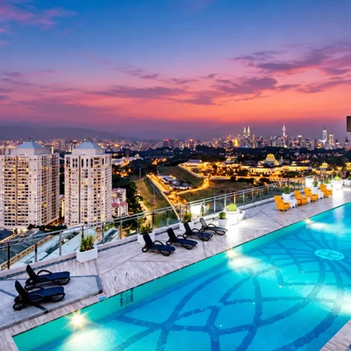 Luxury rooftop infinity pool overlooking the Kuala Lumpur city skyline at sunset, with illuminated high-rise buildings, lounge chairs and vibrant blue pool water.