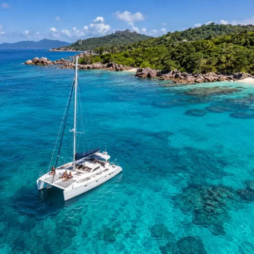 Aerial drone view of a luxury catamaran sailing through clear turquoise Caribbean waters near a lush tropical island.
